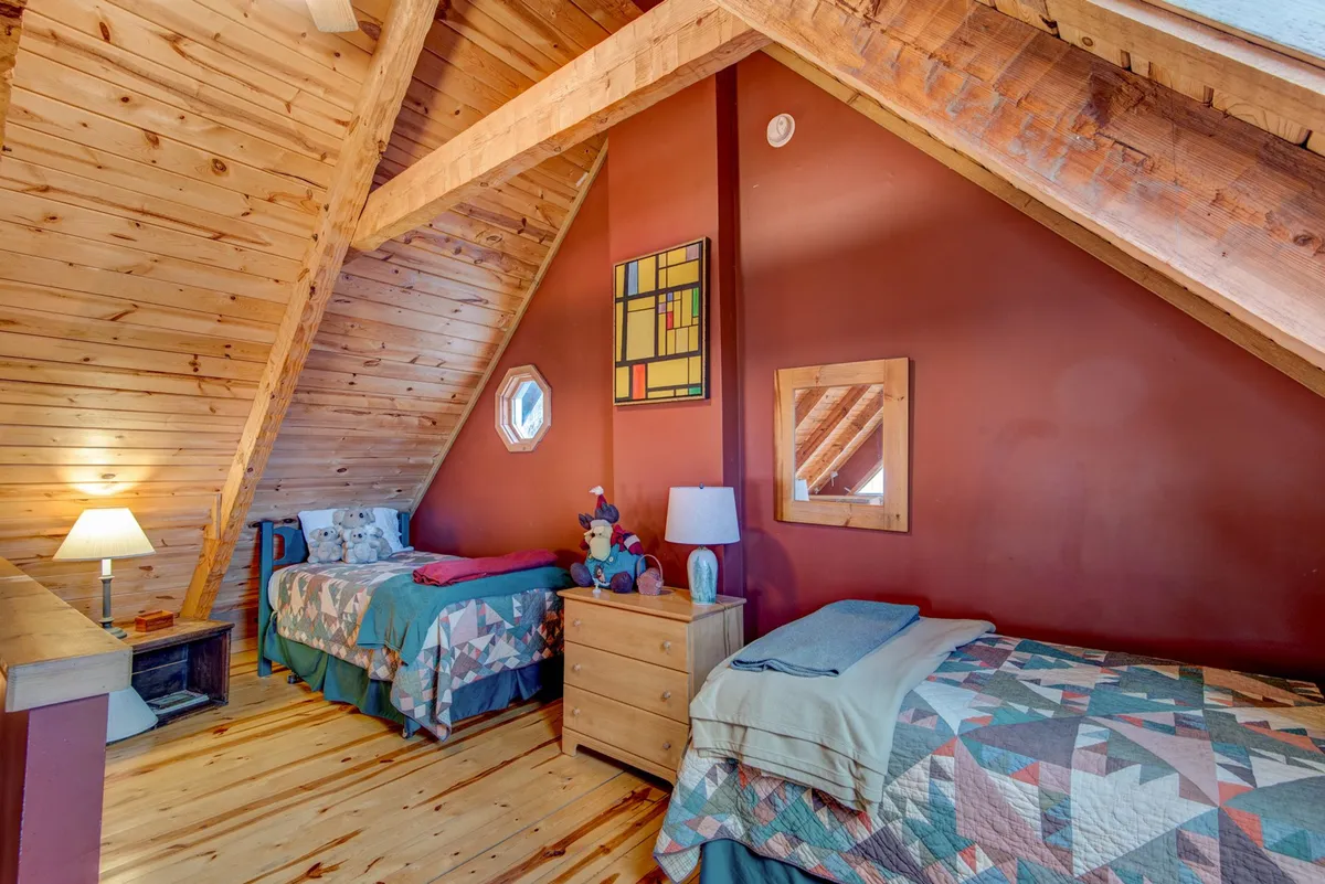 A playful and comfortable sleeping area featuring twin beds with colorful quilted covers, wooden beams overhead, and whimsical wall art. The setup suggests a space ideal for children or extra guests, with a warm, welcoming atmosphere suited for relaxing or playing games.