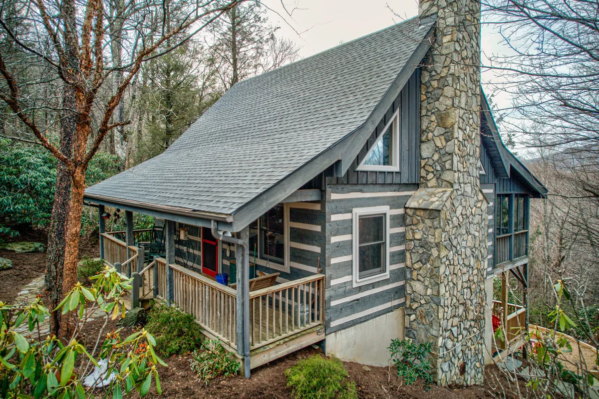 The image shows the exterior of a charming log cabin nestled among trees, featuring a prominent stone chimney and inviting wooden porch. Warm sunset light bathes the wooden siding, evoking a cozy, tranquil atmosphere that's perfect for relaxation. The porch offers space for unwinding and soaking in the natural mountain surroundings.