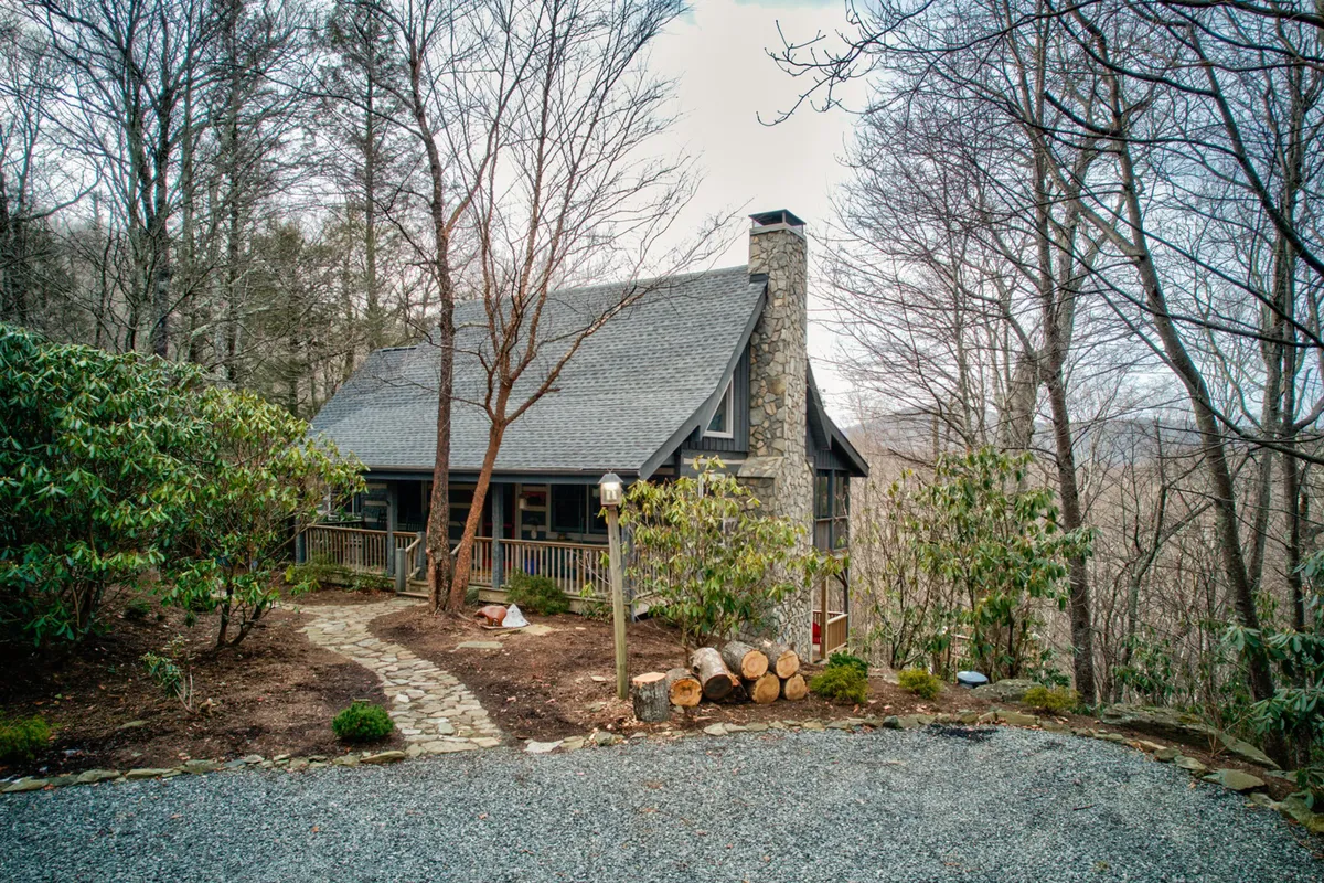 This exterior perspective showcases the house’s stone chimney, the welcoming front pathway, stacked logs, and the scenic wooded backdrop. The view suggests immediate access to trails and natural exploration, with a tranquil, inviting mood that fits the mountain escape theme.