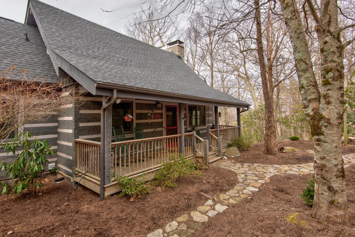 A cozy, classic covered front porch features wooden rocking chairs and a log cabin structure, surrounded by lush trees. The porch offers a welcoming spot for relaxing, reading, or sipping coffee while enjoying the fresh mountain air.