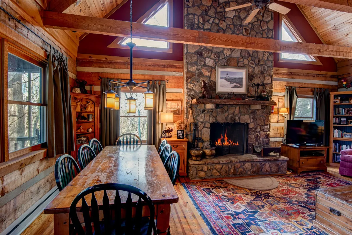 The image focuses on the large, rustic dining table that seats eight, placed beside a stone fireplace and a bookshelf, with ample natural light from large windows. The warm color palette, visible books, and comfortable furniture create an inviting scene for shared meals and relaxation.