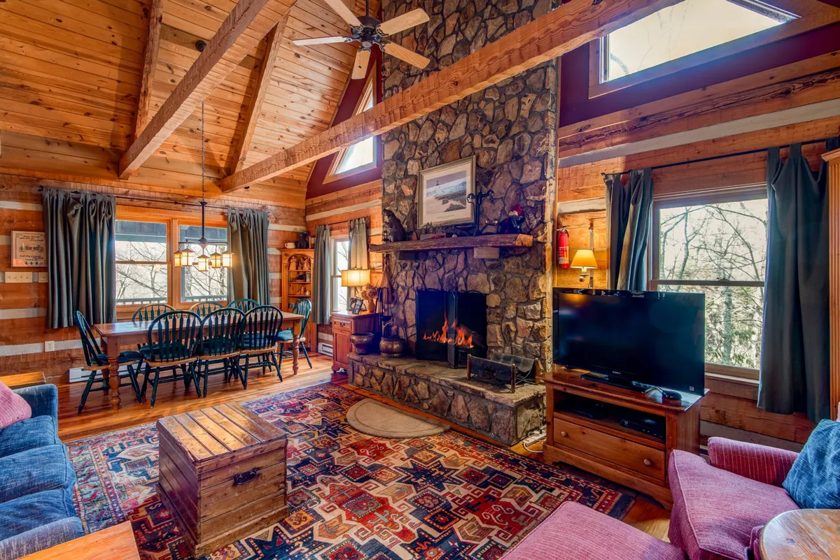This interior image captures a wide view of the great room, showcasing a prominent stone fireplace, a plush rug, and a comfortable sofa. The dining area is visible with a large wooden table, and natural light streams in through windows, highlighting the warm hues of the heart pine floors and the soaring ceiling.