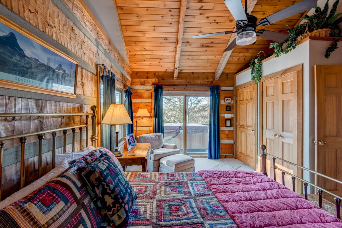 A bright and airy bedroom with a bed covered in a colorful quilt, a large window offering views of the natural surroundings, exposed wooden beams on the ceiling, and a bedside lamp. The room radiates warmth and comfort, inviting guests to wake up to natural light and enjoy quiet moments.