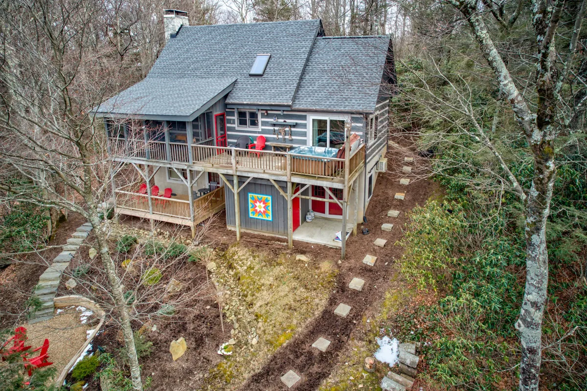 A cozy house with a welcoming porch, red chairs on the deck, and a stone pathway leading up to the entrance. The setting is framed by trees and the soft, natural landscape, suggesting easy access to trails and the outdoors.