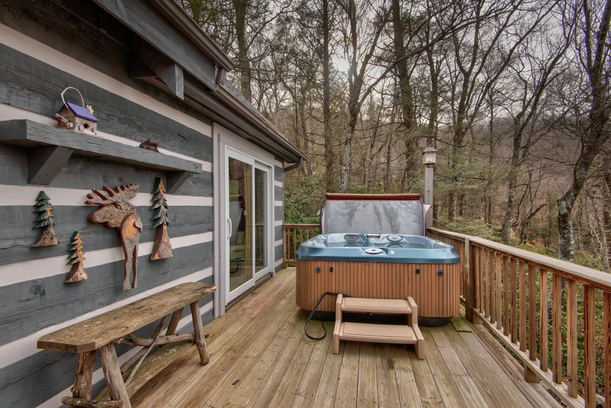 The image shows a cozy backyard scene featuring a hot tub set among trees, accompanied by a wooden bench and decorative wall art. The natural surroundings and inviting hot tub create a retreat-like atmosphere.