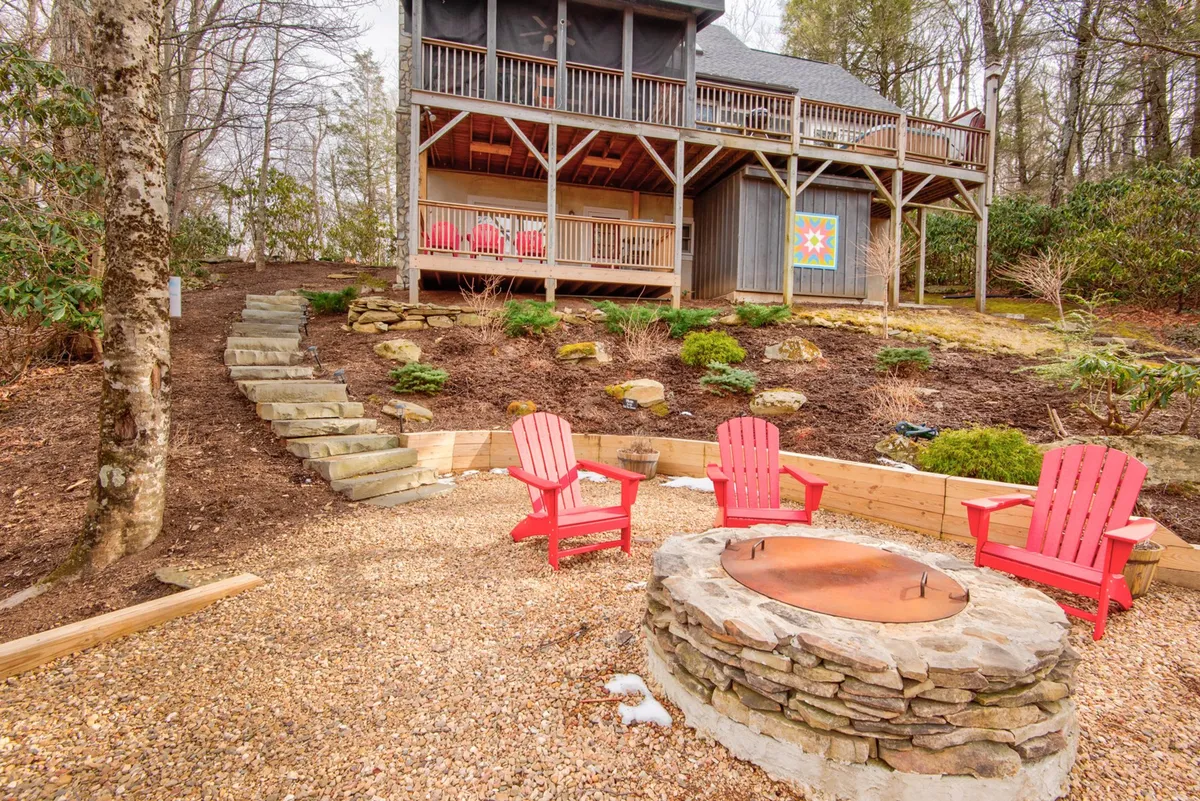 A cozy fire pit sits at the heart of a landscaped garden, surrounded by red Adirondack chairs and the lower deck of the house. The setup is inviting, suggesting evenings spent sharing stories, roasting marshmallows, and enjoying the mountain night air.