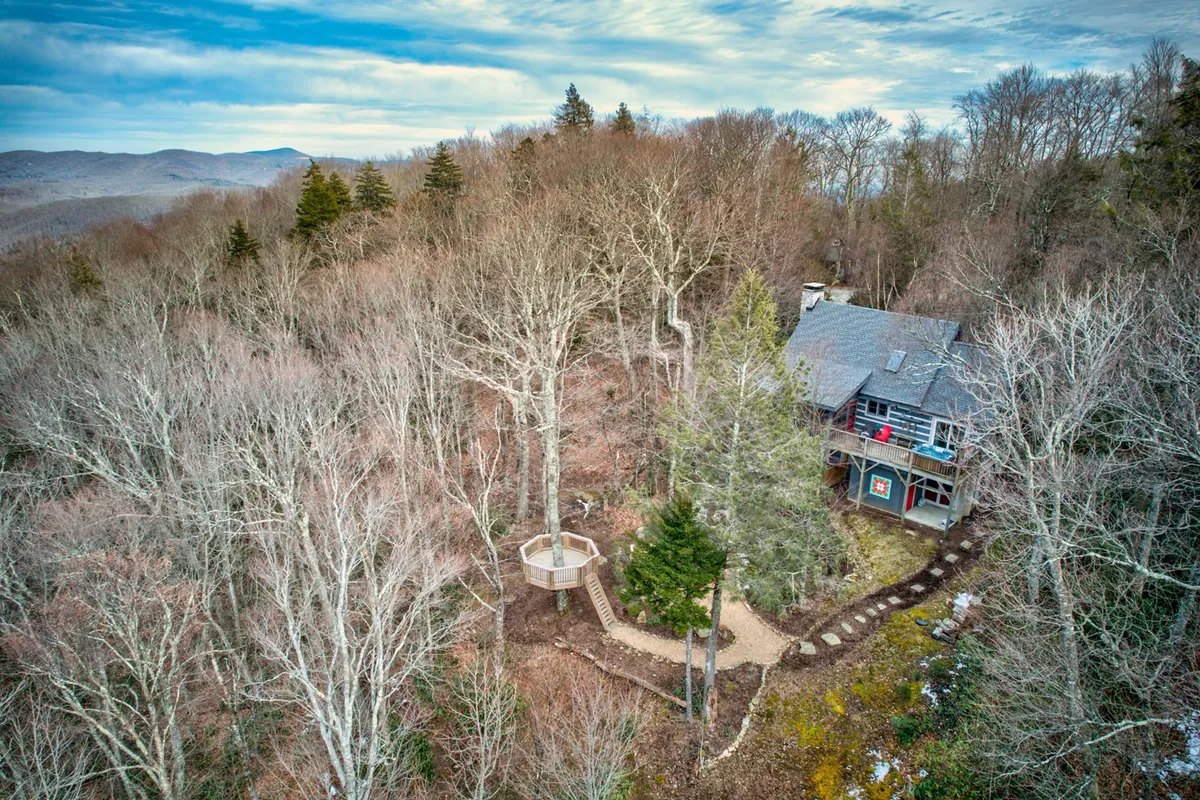 A wide view of the house is framed by lush surrounding trees and a distinctive circular deck. The scene hints at hiking trails and nature walks directly accessible from the property, reinforcing the sense of adventure right outside the door.