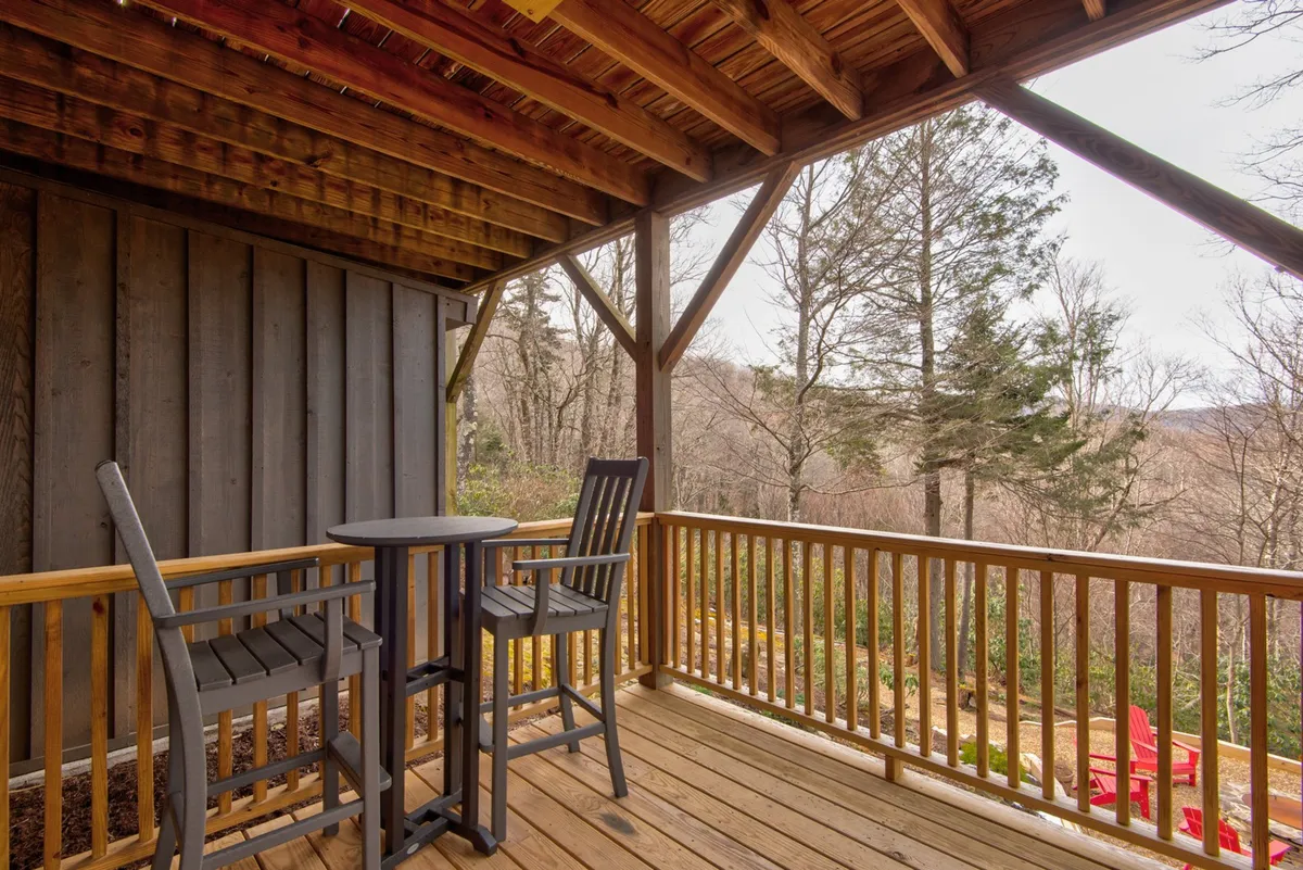 This image displays a wooden deck with a bar-height table and chairs overlooking a natural landscape of trees and open sky. The setting is peaceful and encourages relaxation, reading, or enjoying a drink while surrounded by nature.