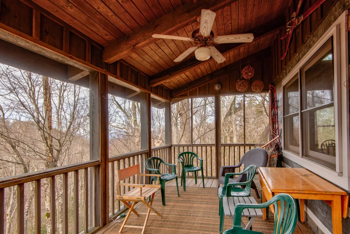 A tranquil screened-in porch featuring a wooden ceiling, green chairs, and a central table. The space is surrounded by screened windows, letting in natural light and views of the outdoors. The setup encourages guests to relax, enjoy a coffee or cocktail, and connect with one another while appreciating the surrounding landscape.